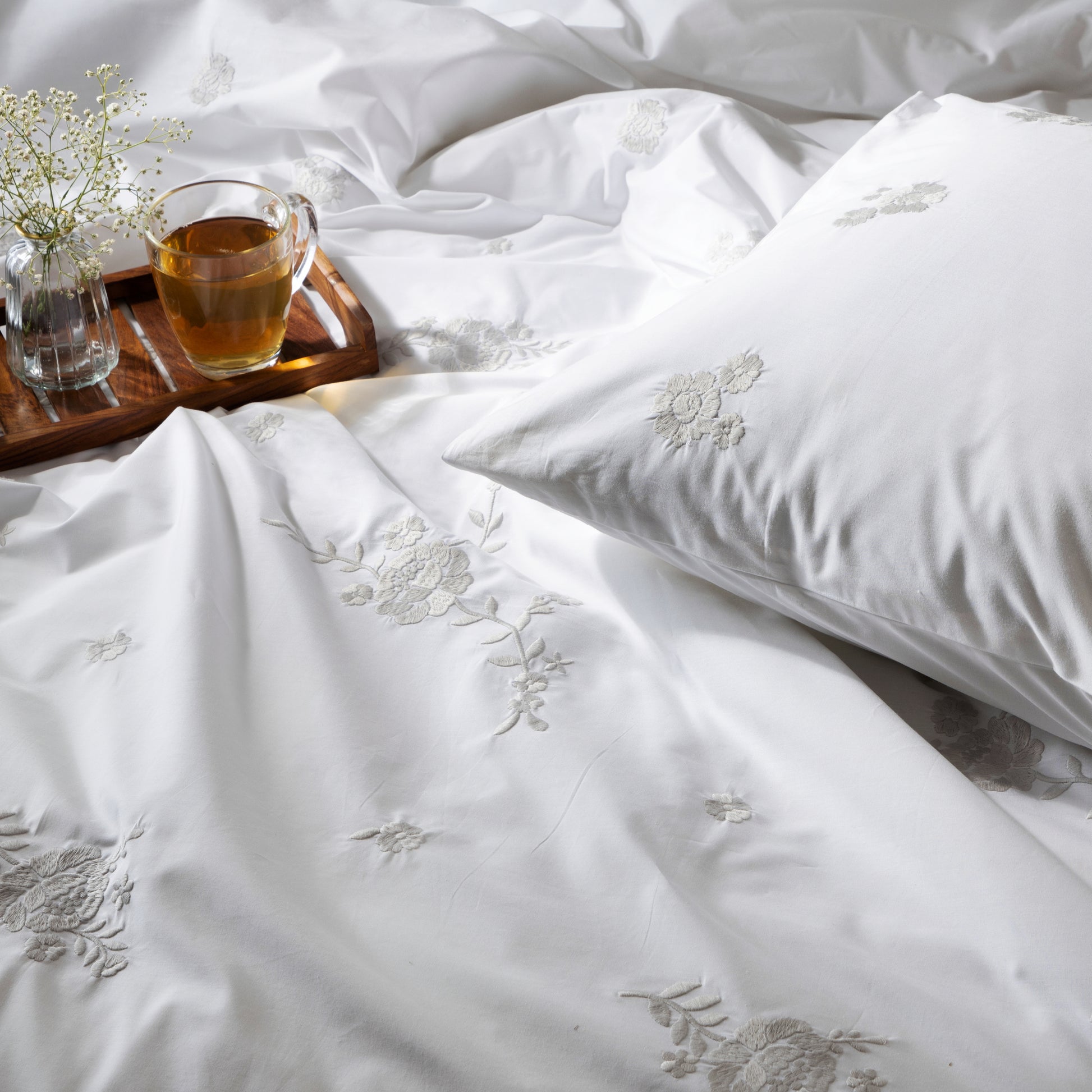 White bedding set with floral embroidery, a cup of tea, and small plant on a wooden tray.