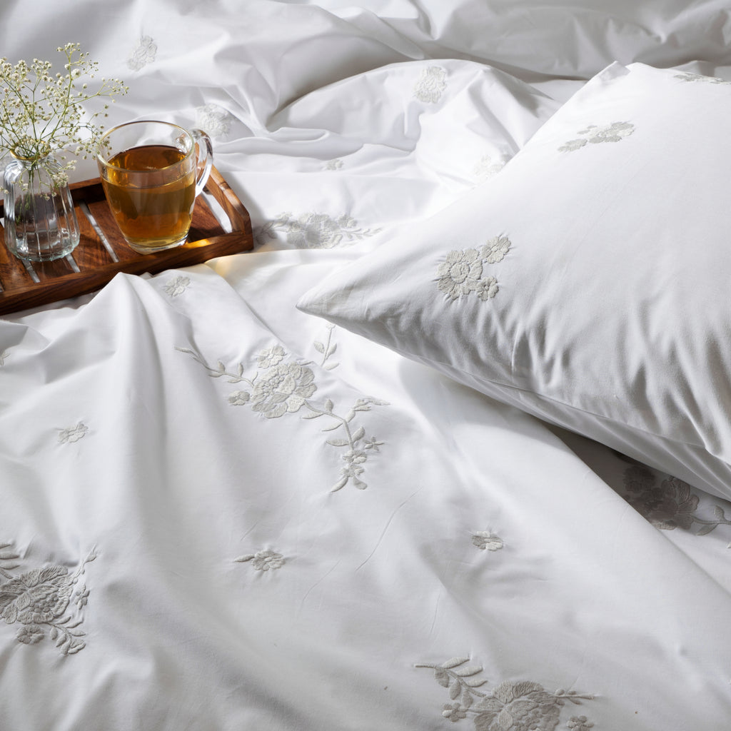 White bedding set with floral embroidery, a cup of tea, and small plant on a wooden tray.